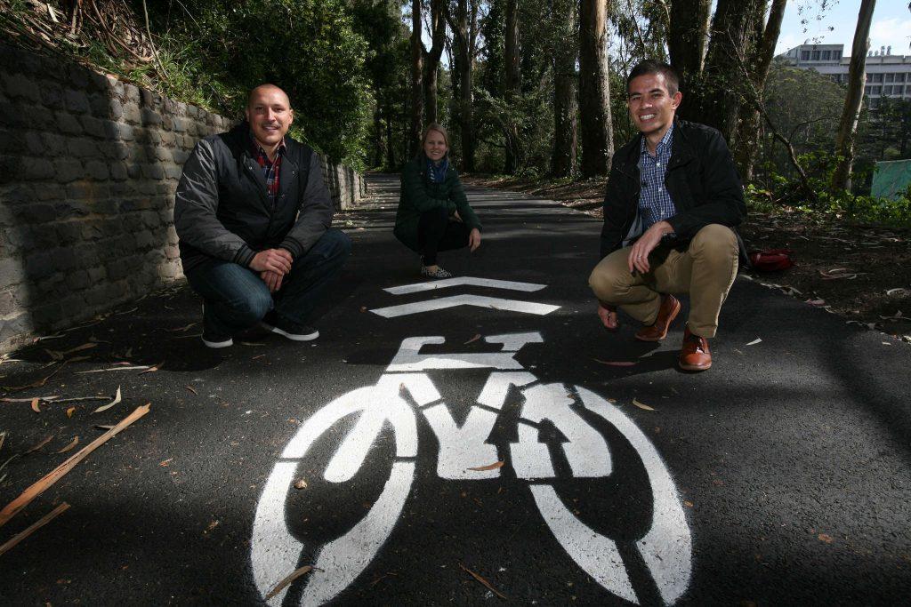 (From right) Nick Kordesch, Caitlin Steele, both are the Sustainability Coordinators at SF State, and Miguel Guerrero, the student coordinator, kneels down next to the newly painted bike path sign which runs along the east side of Cox stadium Monday, March 2. (Daniel Porter / Xpress)