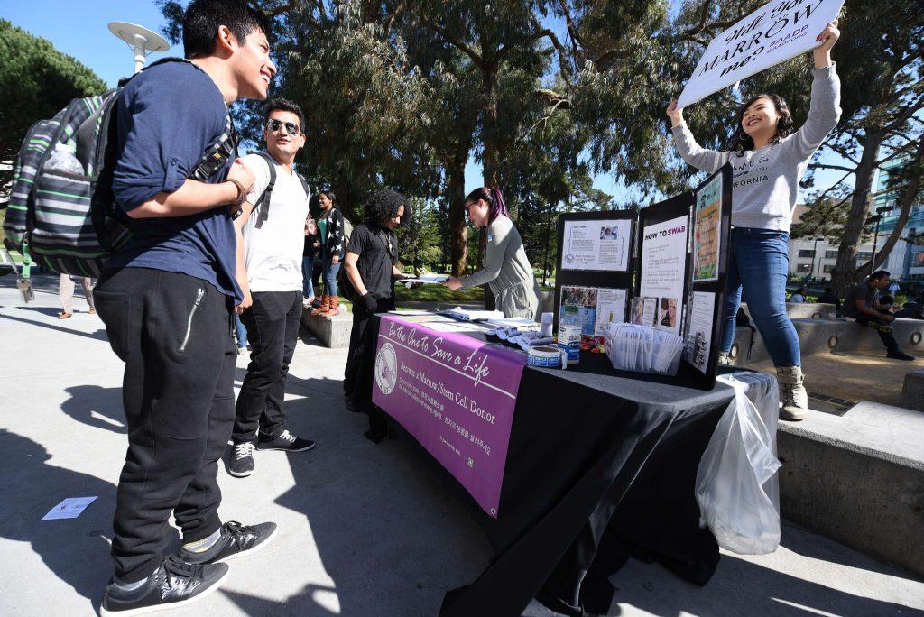 Mauricio Moreno, a junior, smiles at a sign held by Megan Chen saying Marrow me as he visits the AADP table at Malcolm X Plaza Thursday, Feb. 26. (David Henry / Xpress)