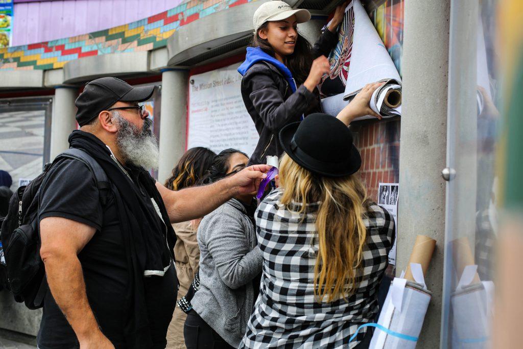 Victor De La Rosa, associate professor at SF State in the Art Department (far left) and his volunteers ( left) Emily Cruz, Veronica Rued ( center) , and Nash Bellows puts up one of his art  pieces at the 16th street Bart station Tuesday, Oct. 27. (Angelica Ekeke/Xpress)