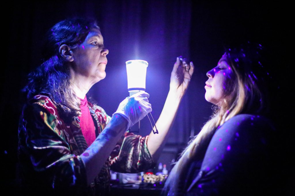 Stacey Dennick owner of Auntie Staceys Face Painting holds a light in front of Carolina Martinezs face durring a face painting session   at the Dia De Los Muertos Fundraising event for All Hands organization at the Raven Bar Wednesday, Nov. 5, 2015. (Angelica Ekeke / Xpress)