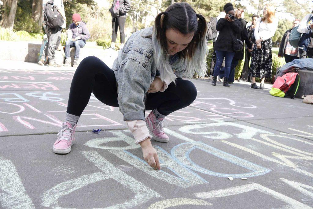 Julia Grace, 22, women and gender studies major, writes in chalk in front of the Wells Fargo and Bank of America ATM machines during the protest against the DAPL and KPL funding from the two banks. (Mason Rockfellow/Xpress)