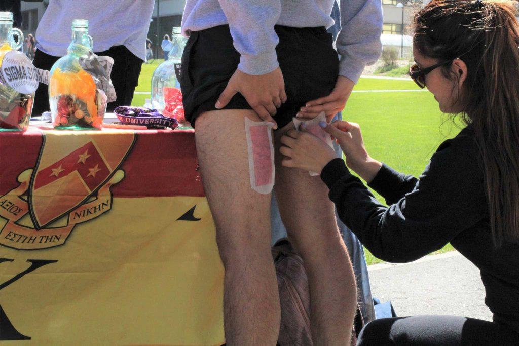 Edgar Avila (left), from Phi Kappa Tau holds still as Corrina Guerrero (right), from Phi Sigma Sigma prepares a quick hair removal on the area of choice on March 29, 2017 at SF State. This is one of the many games Phi Kappa Tau is hosting to raise money for a childrens camp in San Francisco, Calif. (Alina Castillo/Xpress)
