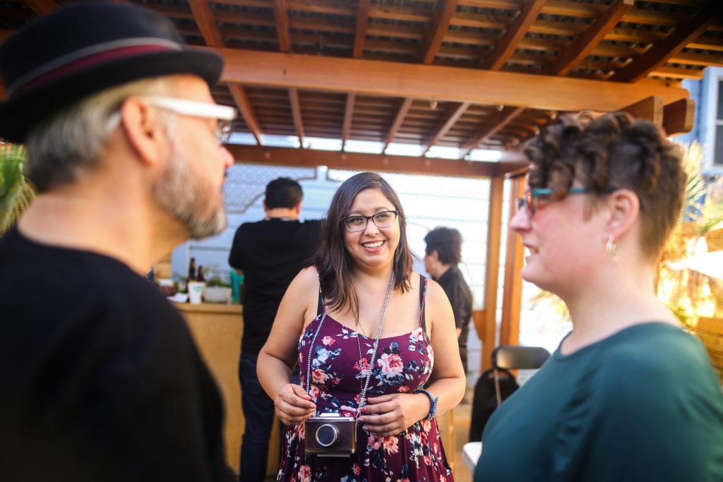 El Tecolote volunteer photographer Drago Renteria (left) and his girlfriend Mantle, chat with Mabel Jimenéz-Hernandez during Hernandezs going away party at Acción Latina in San Francisco on Saturday, August 25, 2018. (Adelyna Tirado/Golden Gate Xpress)