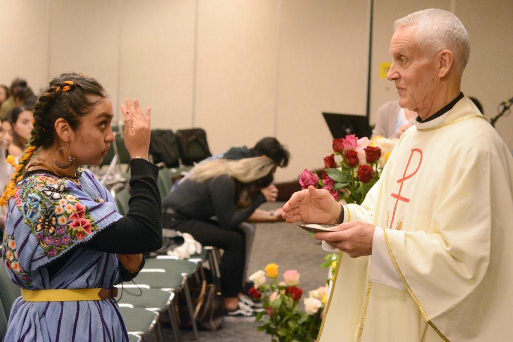 Angie Arroyo (left), 20, a third-year child development major with an emphasis in youth and adult school work makes the sign of the cross while Father Rick Van De Water (right), 68, Chaplain for Arabic-speaking members of the Roman Catholic Archdiocese from St. Thomas More Church gives communion at a Catholic mass for Lady Guadalupe at SF State on Wednesday, Dec. 12, 2018. (Aaron Levy-Wolins/Golden Gate Xpress)