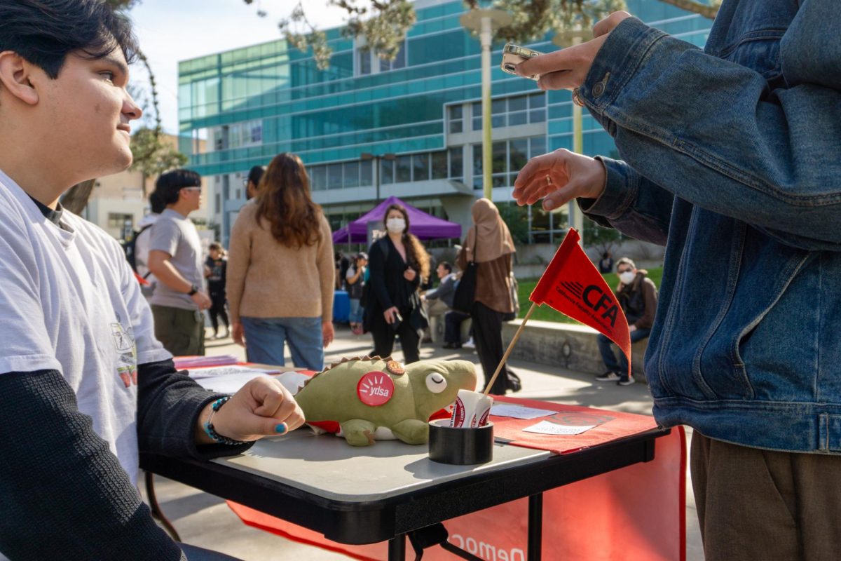 A plush alligator and a California Faculty Association flag are seen between students interacting at the Young Democratic Socialists of America table at Malcolm X Plaza on Monday, Jan. 29, 2024. (Dan Hernandez / Golden Gate Xpress)