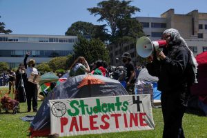 A protestor directs chants through a megaphone as other protestors set up tents at San Francisco State Universitys quad on Monday, April 29, 2024. (Neal Wong / Golden Gate Xpress)