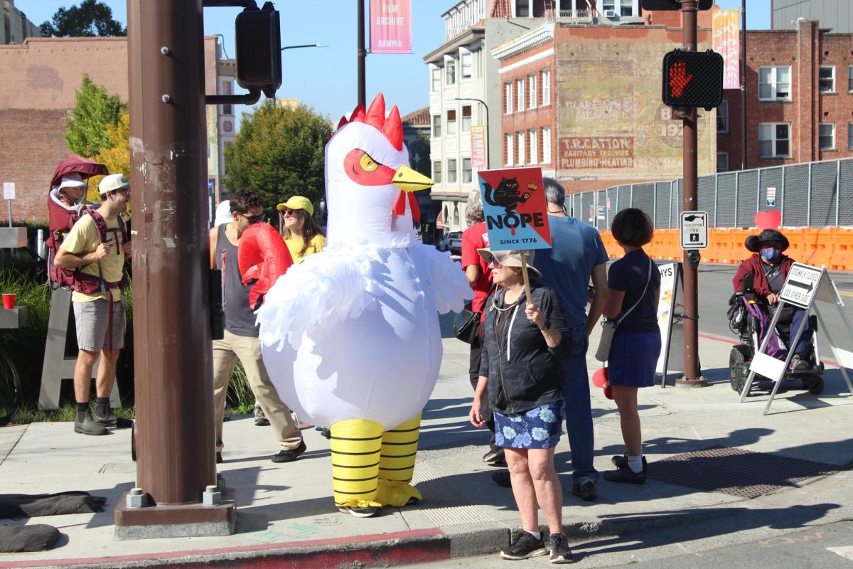 A protester holds a sign saying “Nope Since 1776” beside a person wearing an inflatable chicken costume on the intersection of Addison and Oxford streets on Saturday, Oct. 18, 2025. (Paul Singh / Golden Gate Xpress)